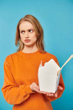 Turuncu süveterli şık bir kadın zarif bir şekilde nefis bir yemek kutusu tutuyor, moda ve gastronominin karışımını gösteriyor..