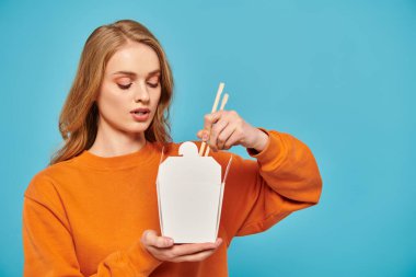 A stylish woman in an orange sweater gracefully holds box of Asian food in her hand. clipart