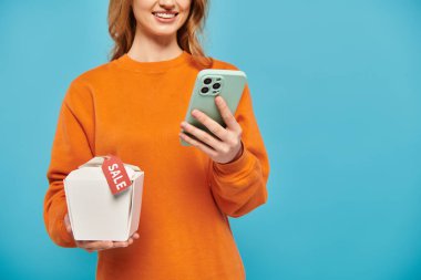 Elinde cep telefonu ve bir kutu Asya yemeği olan sarışın kadın, modern yemek deneyiminin tadını çıkarıyor..