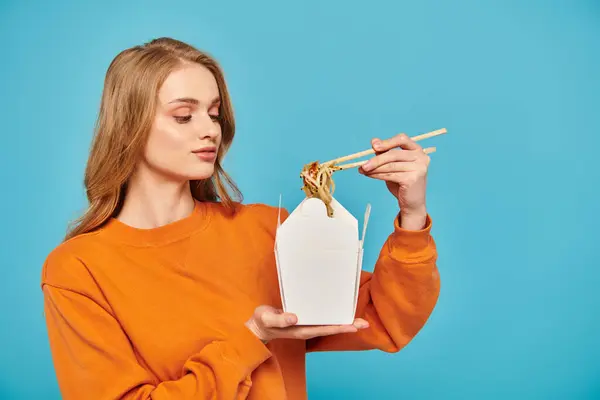 一位穿着橙色毛衣的金发女人拿着一个盛满面条的白色容器 用筷子展示亚洲菜 — 图库照片