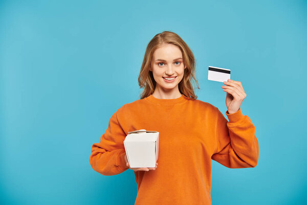 An elegant woman appears content as she holds a credit card in one hand and a food box in the other, symbolizing online shopping.