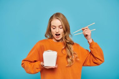 Güzel sarışın bir kadın elinde bir kutu erişte ve yemek çubuğu tutuyor. Zarafet saçıyor ve Asya mutfağından zevk alıyor..