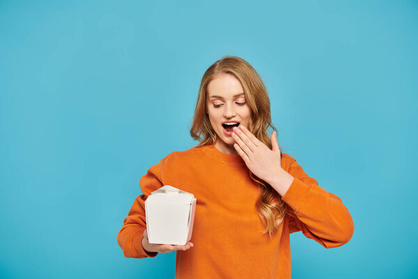 A woman with blonde hair holding a box of Asian food in front of her face.