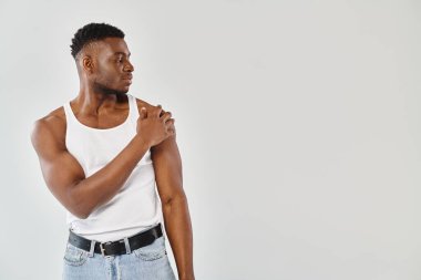 Beyaz kolsuz tişörtlü ve kot pantolonlu genç, seksi bir Afro-Amerikalı, gri bir arka plana karşı stüdyoda güven içinde poz veriyor..