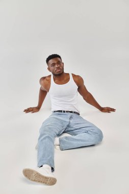 Genç ve seksi Afrikalı Amerikalı bir adam kollarını bir stüdyoda gri bir arka plana uzatarak yerde oturuyor..