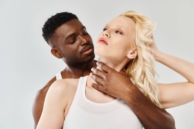 Beyaz kolsuz bluzlu bir kadının yanında bir adam duruyor. Stüdyoda, arka planı gri olan bir kadın..