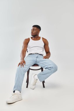 Beyaz kolsuz tişörtlü genç bir adam gri arka planı olan bir stüdyoda sandalyede oturuyor..