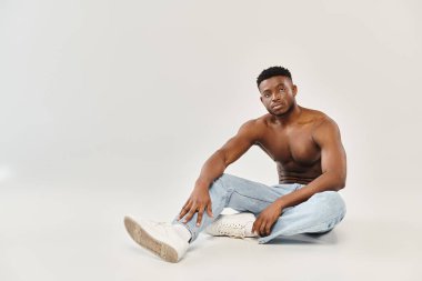 Gri arkaplanı olan bir stüdyoda yerde oturan genç, Afro-Amerikan bir adam..