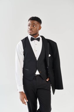 Genç ve yakışıklı bir Afrikalı Amerikalı damat, gri bir stüdyo geçmişine karşı şık bir smokin içinde kendine güvenen bir poz veriyor..