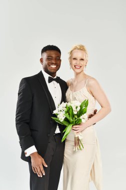 Gelinlik giymiş güzel bir sarışın gelin ve smokinli bir Afro-Amerikan damat gri bir stüdyo arka planında zarif bir şekilde duruyorlar..
