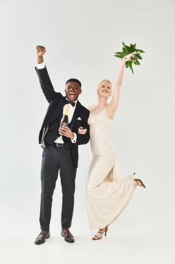 Gelinlik giymiş güzel bir sarışın gelin ve gri arka planda bir stüdyoda çiçekleri tutan Afrikalı bir damat..