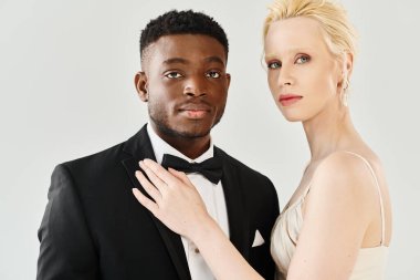 A beautiful blonde bride in a white dress stands next to an African American groom in a tuxedo, exuding elegance and love. clipart