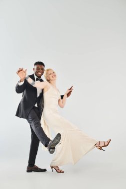 Gelinlik giymiş güzel bir sarışın gelin ve gri arka planda zarif bir şekilde dans eden Afro-Amerikan damat..