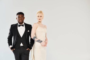 An African American man in a tuxedo stands next to a beautiful blonde woman in a white dress in a studio against a grey background. clipart