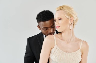 Gelinlik giymiş güzel bir sarışın gelin ve smokinli bir Afro-Amerikan damat, gri bir arka planda zarif bir şekilde stüdyoda duruyorlar..