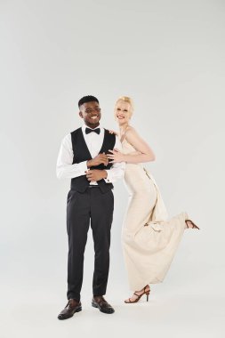 Gelinlik giymiş güzel bir sarışın gelin ve smokinli Afrikalı bir damat gri bir arka planda stüdyoda birlikte poz veriyorlar..