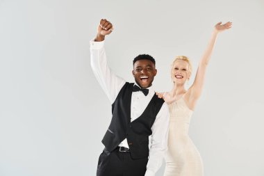 Gelinlik giymiş güzel bir sarışın gelin ve Afrikalı bir damat gri bir arka planda zarif bir şekilde stüdyoda poz veriyorlar..