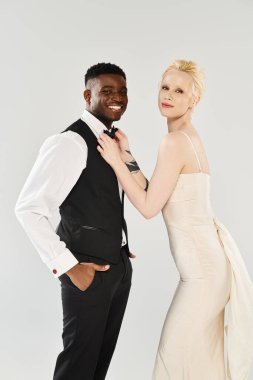 Gelinlik giymiş güzel bir sarışın gelin, gri arka plandaki bir stüdyoda Afrikalı Amerikalı bir damadın yanında duruyor..