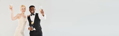 Gelinlik giymiş güzel bir sarışın gelin ve gri arka planda bir stüdyoda şampanya kadehleri tutan Afrikalı bir damat..