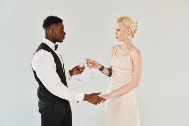 Gelinlik giymiş sarışın gelin ve Afrikalı Amerikalı damat gri arka planda romantik bir anda şampanya kadehlerini tokuşturuyorlar..
