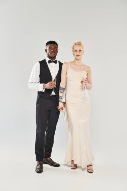 Gelinlik giymiş güzel bir sarışın gelin ve smokinli bir Afrikalı Amerikalı damat gri bir arka planda stüdyoda yan yana duruyorlar..