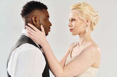 Gelinlik giymiş güzel bir sarışın gelin ve gri arka planda bir stüdyoda birlikte duran Afrikalı Amerikalı damat..