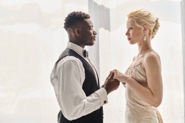 A beautiful blonde bride in a wedding dress and an African American groom standing together in a studio, on a grey background. clipart