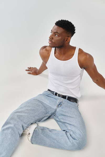 A stylish African American man relaxes on the ground, clad in denim jeans against a grey backdrop.