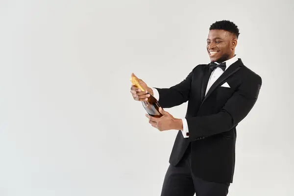 stock image Handsome African American groom in a tuxedo holding a bottle of champagne, exuding elegance and sophistication on a grey studio background.