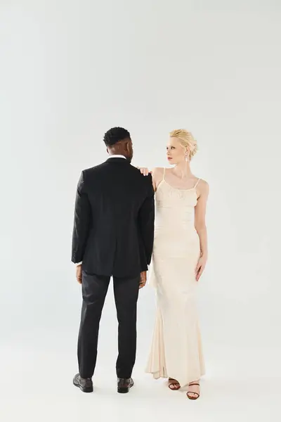 stock image A beautiful blonde bride in a wedding dress and an African American groom stand next to each other in a studio on a grey background.