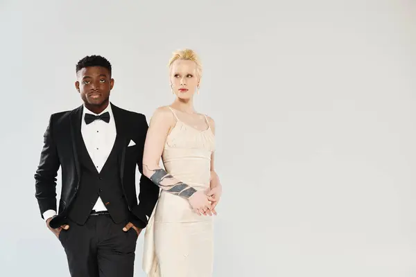 Stock image An African American man in a tuxedo stands next to a beautiful blonde woman in a white dress in a studio against a grey background.