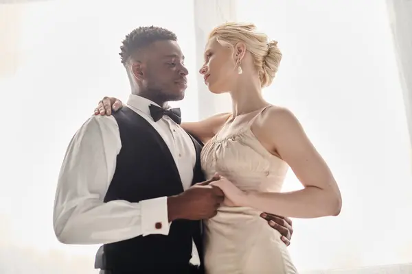 stock image A beautiful blonde bride in a wedding dress stands next to an African American groom in a tuxedo against a grey studio background.