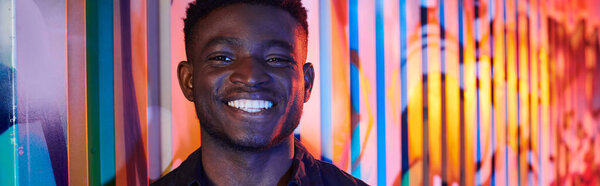 A man stands boldly in front of a vibrant, colorful wall on an urban street filled with graffiti.
