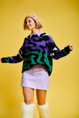 Parlak kazaklı, kısa saçlı, neşeli bir kadın sarı arka planda etkin bir şekilde poz veriyor.