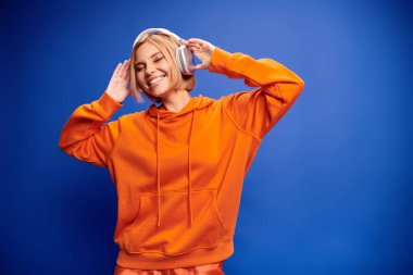 Kısa sarı saçları ve kulaklıkları olan şık bir kadın mavi arka planda müziğin keyfini çıkarıyor.