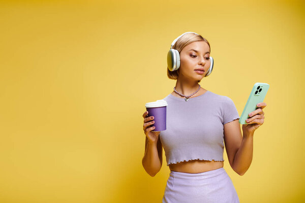 appealing cheerful woman with headphones and cell phone enjoying her coffee on yellow backdrop