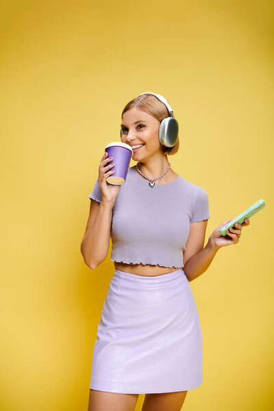 beautiful cheerful woman with headphones and cell phone enjoying her coffee on yellow backdrop