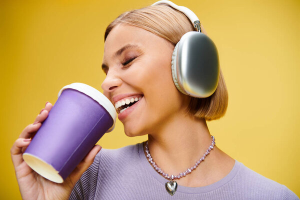 joyous stylish woman with headphones enjoying music and hot coffee while posing on yellow backdrop