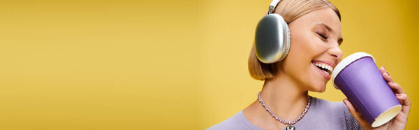 happy woman with headphones enjoying music and hot coffee while posing on yellow backdrop, banner