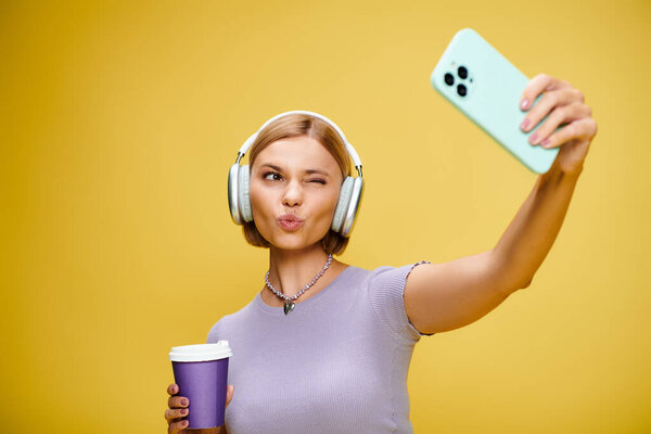 alluring cheerful woman with headphones and cell phone enjoying her coffee on yellow backdrop