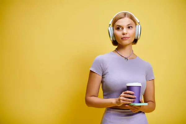 黄色い背景に彼女のコーヒーを楽しんでいるヘッドフォンと携帯電話を持つ若い陽気な女性 — ストック写真