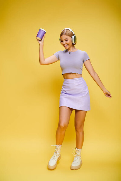 glad trendy woman with headphones enjoying music and hot coffee while posing on yellow backdrop