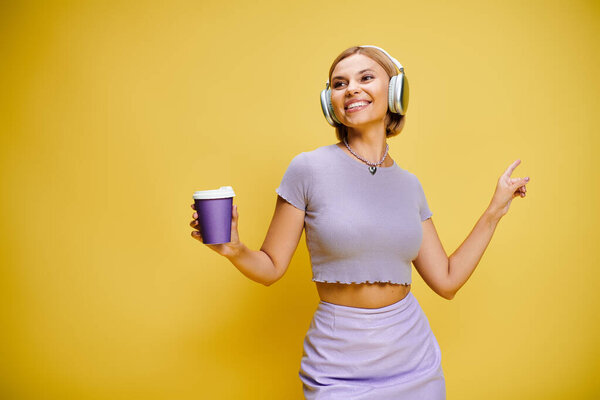 cheerful trendy woman with headphones enjoying music and hot coffee while posing on yellow backdrop