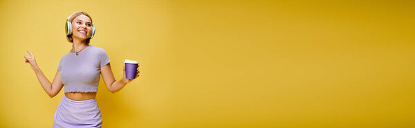 merry woman with headphones enjoying music and hot coffee while posing on yellow backdrop, banner
