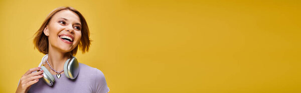 young joyous woman with short blonde hair and headphones enjoying music on yellow backdrop, banner