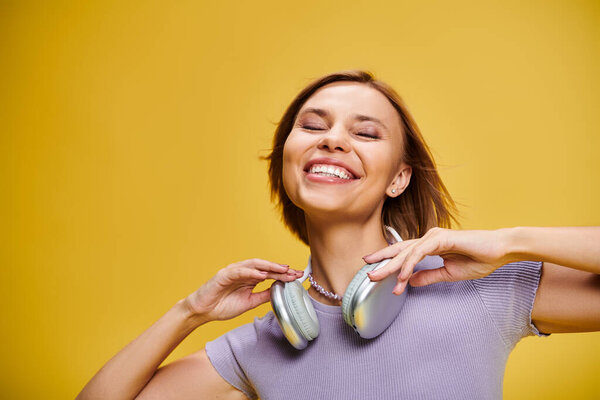 debonair joyous woman with short blonde hair and headphones enjoying music on yellow backdrop