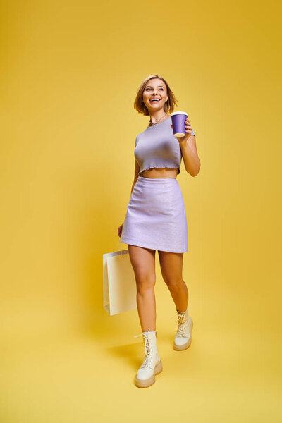 joyful young woman in vibrant attire with short hair posing with coffee and shopping bag in hands