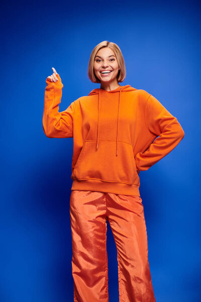 attractive cheerful woman with short hair in vibrant orange hoodie posing actively on blue backdrop
