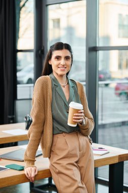 Bir kadın, şirket ofisindeki bir masanın önünde elinde bir fincan kahveyle rahatça duruyor..