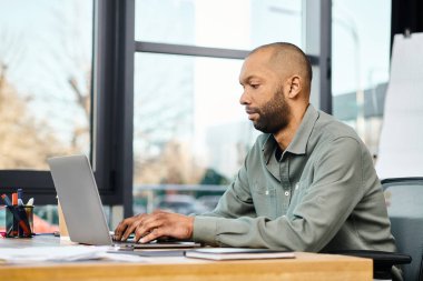 Şirket ofisindeki bir adam, bir proje üzerinde çalışırken yoğun bir şekilde laptopuna odaklanmış bir şekilde masasında oturuyor..
